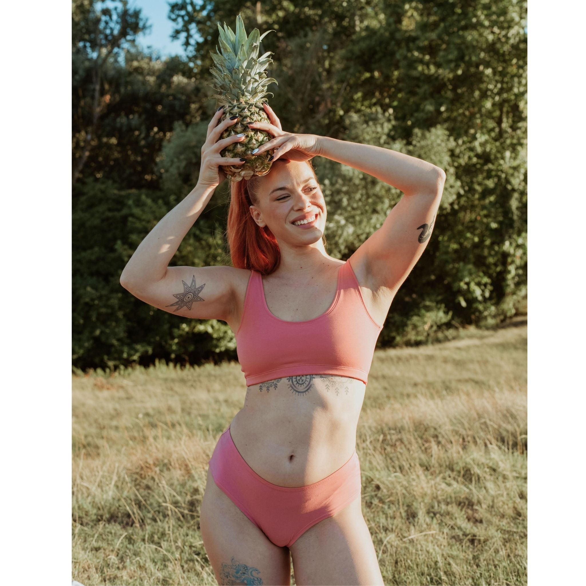 Woman wearing Desert Rose Organic Cotton Hipster Panty Panty holding pineapple on her head in a sunlit outdoor setting.